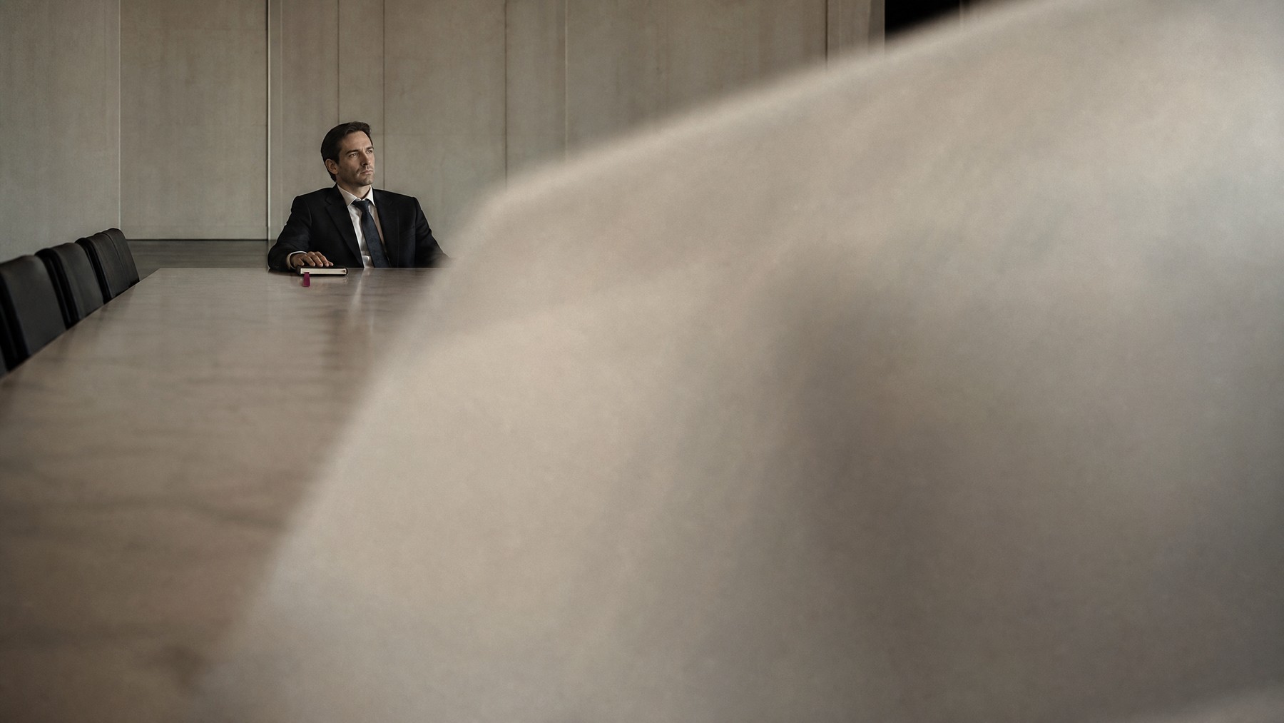 A solitary founder sits at the far end of a long boardroom table while the foreground falls softly out of focus.